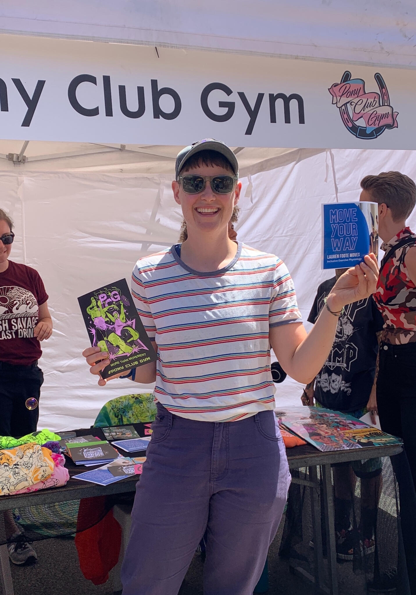 Lauren is wearing a rainbow striped t shirt and holding flyers while smiling in the sun.